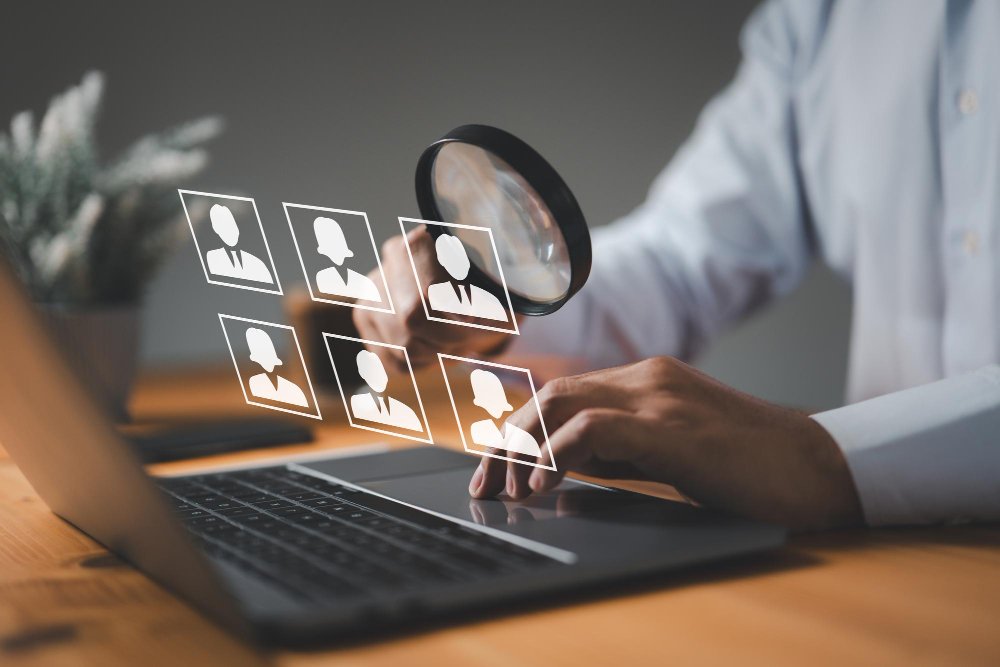 Person holding a magnifying glass over virtual candidate profiles on a laptop